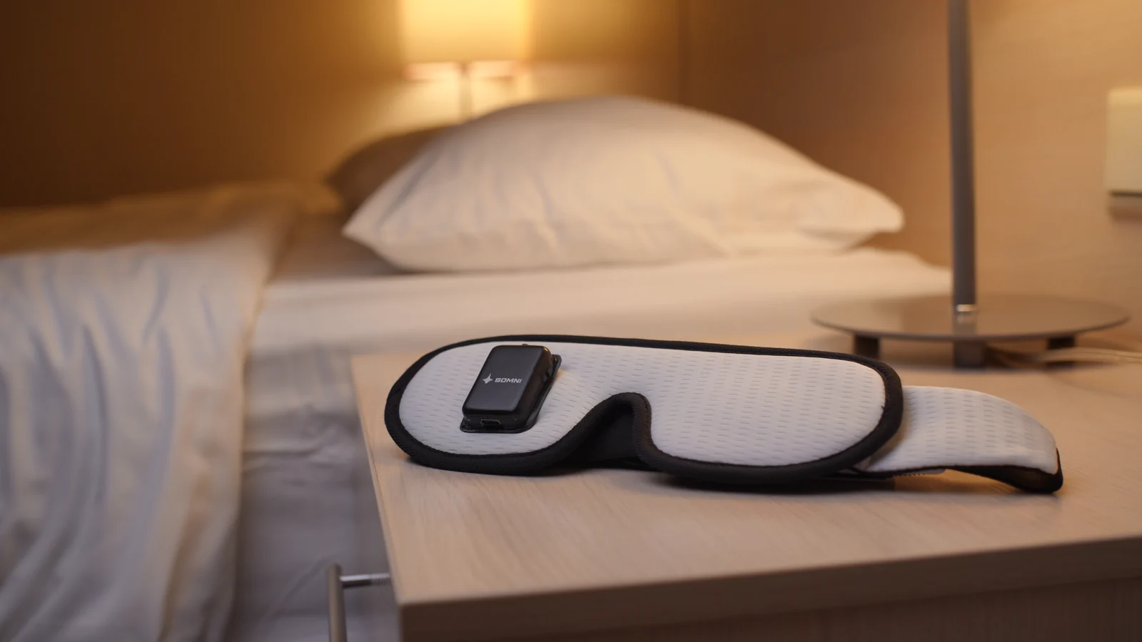 Somni Mask on a bedside table beside a bed and lamp